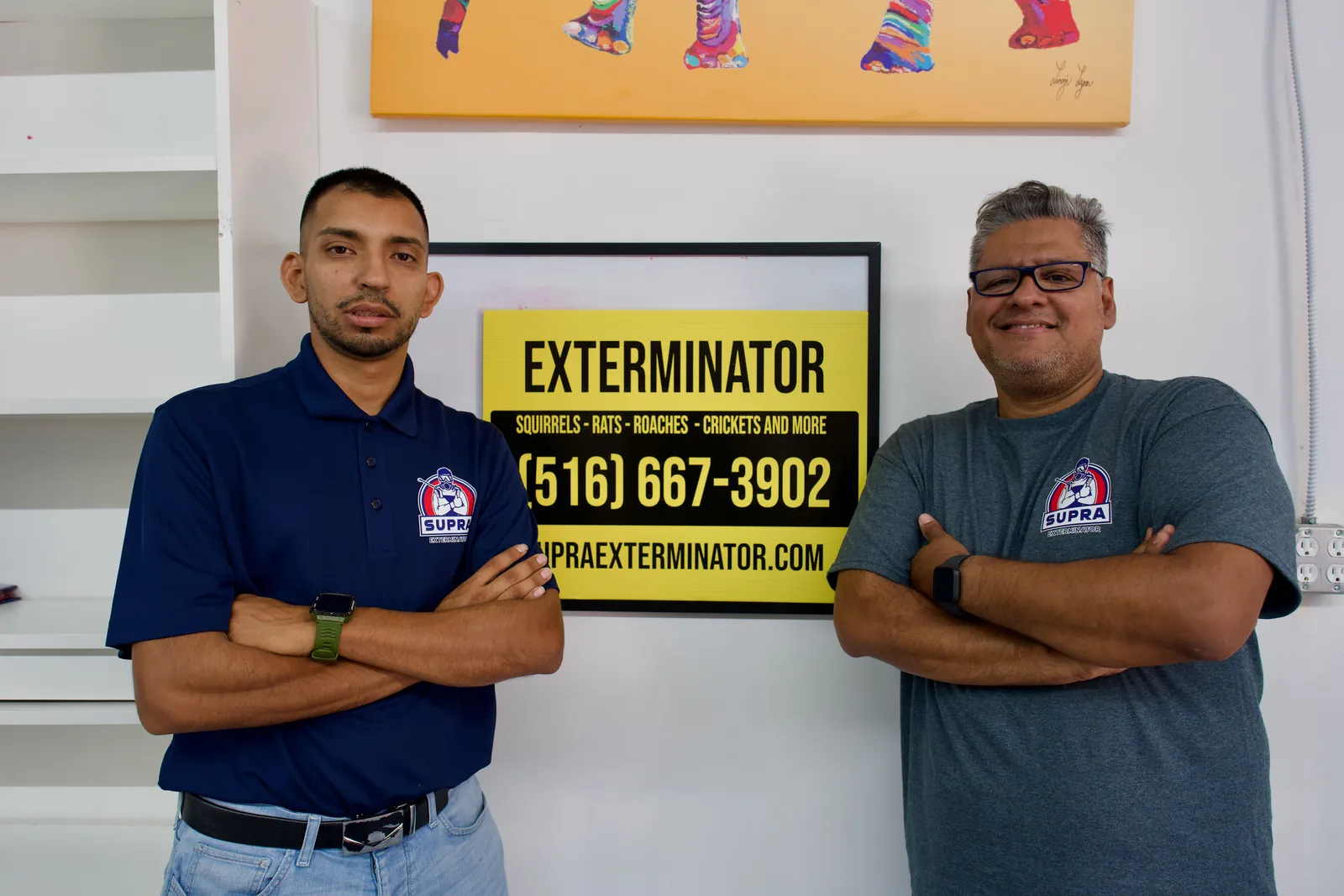 Two Supra Exterminator owners standing beside an exterminator sign showing the phone number (516) 667-3902